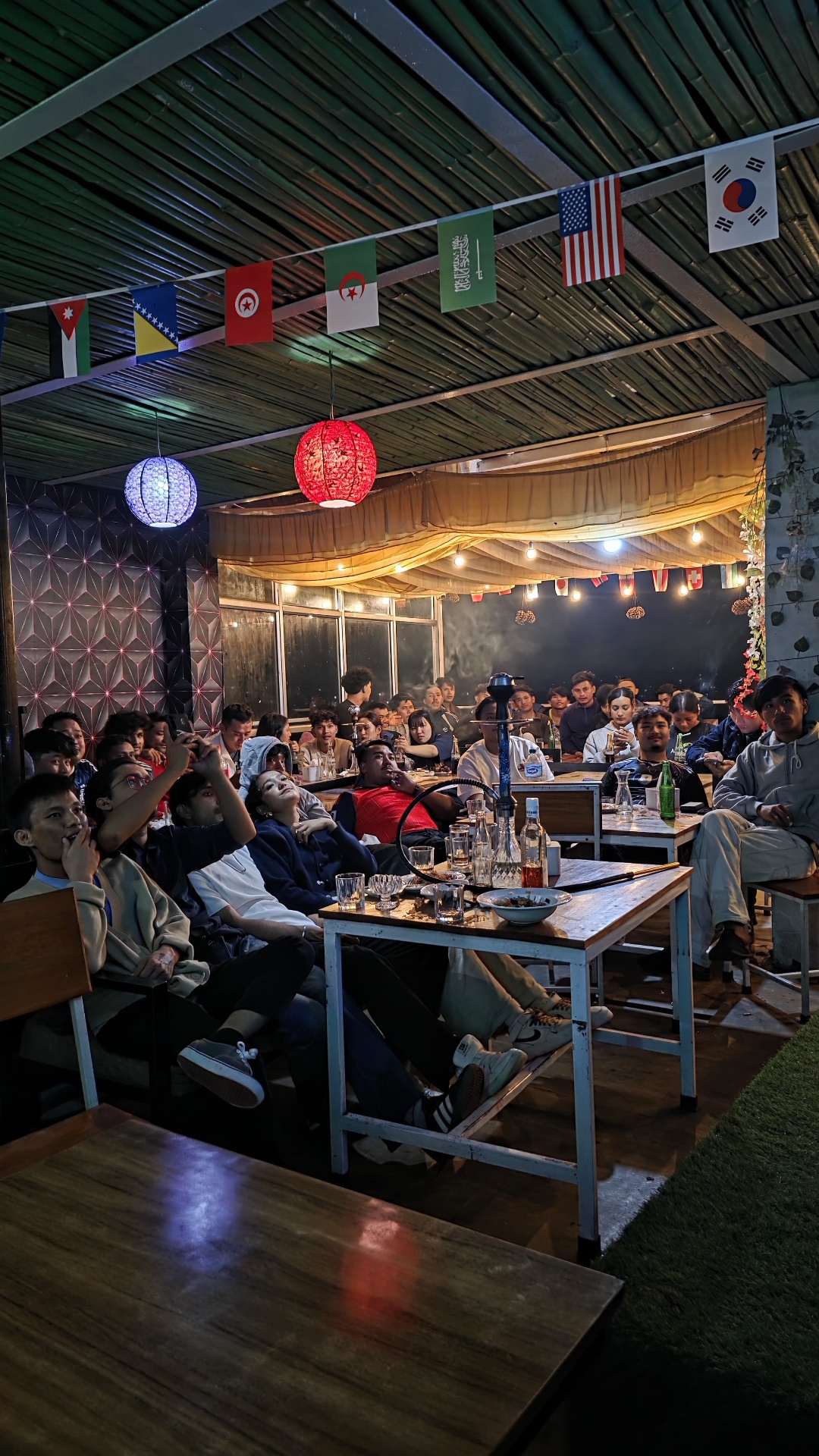 A full house of guests watching a match together on the covered rooftop with lanterns and flags overhead